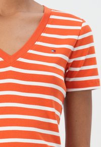 Oransje og hvit stripe V-hals t-skjorte, med korte ermer og et lite logo-patch, laget av myk stoff med en jevn tekstur.