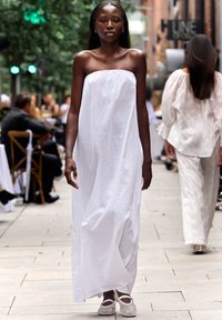 Hvit, stroppeløs maxikjole i lett stoff, med flytende silhuett, brukt med beige fottøy. Modellen går på en fortau kantet med grønne planter.