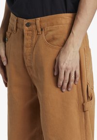Pantalones cargo de algodón marrón con un corte clásico, que cuentan con bolsillos laterales y una tela texturizada. La cintura incluye un cierre de botón.