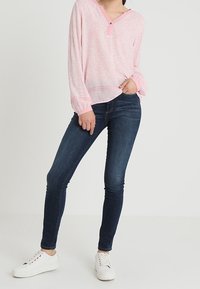Maglietta a maniche lunghe rosa chiaro con scollo a V e chiusura con un solo bottone, abbinata a jeans skinny blu scuro e sneakers bianche.