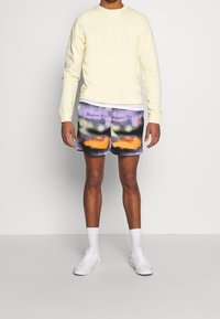Felpa gialla pastello da uomo, pantaloni corti viola tie-dye con motivi arancioni e neri, e sneakers bianche. Design semplice con vestibilità casual.