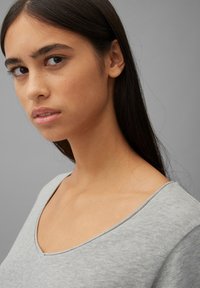 Grå v-hals t-skjorte med korte ermer, myk tekstur og minimal sømdetalj langs halslinjen. Modellen poserer med et nøytralt uttrykk.