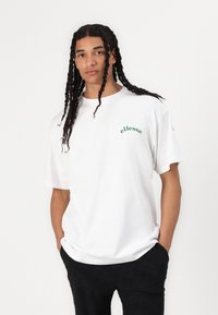 Camiseta de algodón blanca con cuello redondo y mangas cortas. Presenta texto de logo verde en el pecho. Corte holgado, combinada con pantalones oscuros.