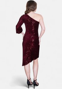 Robe en sequins bordeaux avec une manches longue et une épaule dénudée ; ourlet asymétrique ; associée à des chaussures à talons noirs.