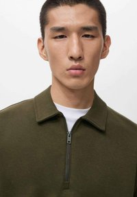 Olive zelen zip-up dukserica z mandarin ovratnikom, z gladko teksturo in kratkim sprednjim zipperjem. Nosi se preko bele majice.