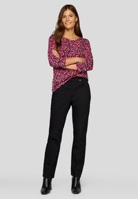 Frau mit verschränkten Armen, die einen rot-schwarz gemusterten Langarmshirt, schwarze Hose und schwarze Stiefeletten vor einem einfarbigen Hintergrund trägt.