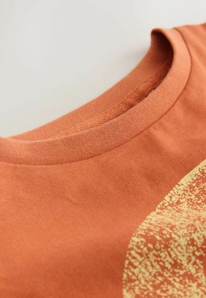 Rozsda színű kerek nyakú póló közeli kép a nyakrészről és a részleges sárga texturált kör alakú mintáról a szöveten.