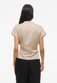 Blouse en satin beige à manches courtes, avec un léger éclat, une silhouette ajustée et une texture lisse. Aucun motif ni élément métallique visible.