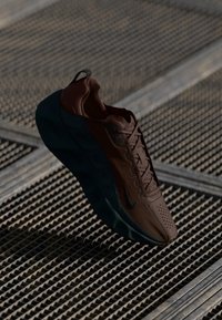 Chaussure de sport marron avec une surface texturée, des accents noirs et une semelle arrondie. Présente un design à lacets et une languette au niveau du talon.