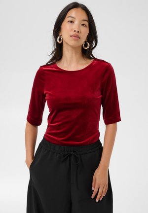 Femme aux longs cheveux foncés portant des boucles d’oreilles argentées en forme de créoles, un haut rouge en velours à manches courtes et un pantalon noir, les mains dans les poches.