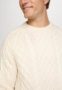 Cremefarbener Kabelstrickpullover mit rundem Ausschnitt, der strukturierte Muster und gerippte Bündchen aufweist. Der Stoff wirkt weich und warm.