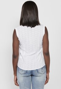 Débardeur en coton blanc sans manches avec broderie œillet, encolure ronde et ourlet arrondi. Associé à un jean en denim bleu.
