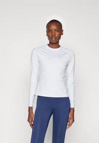 Lys grå langermet treningstopp med rund hals. Glatt, elastisk stoff med en subtil logo på brystet. Kombinert med marineblå tights.
