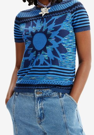 Haut en maille bleu à manches courtes à motif avec manches rayées et motif en forme de rayons de soleil, porté avec un jean taille haute bleu clair, mains dans les poches.