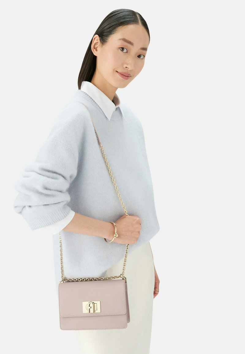 Borsa di pelle viola chiaro con una chiusura e una tracolla in tono oro, dalla texture liscia, portata da una persona che indossa un maglione azzurro chiaro.