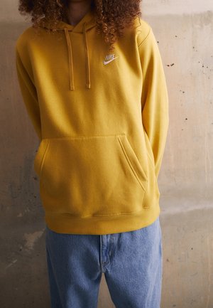 Person iført en sennepsgul Nike-hoodie med forlomme og lyseblå jeans, stående foran en tekstureret væg.