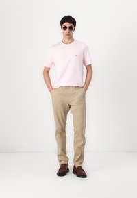 T-shirt rose clair avec un petit logo, pantalon beige et chaussures brunes à enfiler. Modèle portant des lunettes de soleil rondes, se tenant devant un fond blanc.