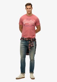 Camiseta de algodón rojo jaspeado con el logo "SuperDry", jeans azul claro, zapatillas color crema y una camisa de cuadros anudada a la cintura.