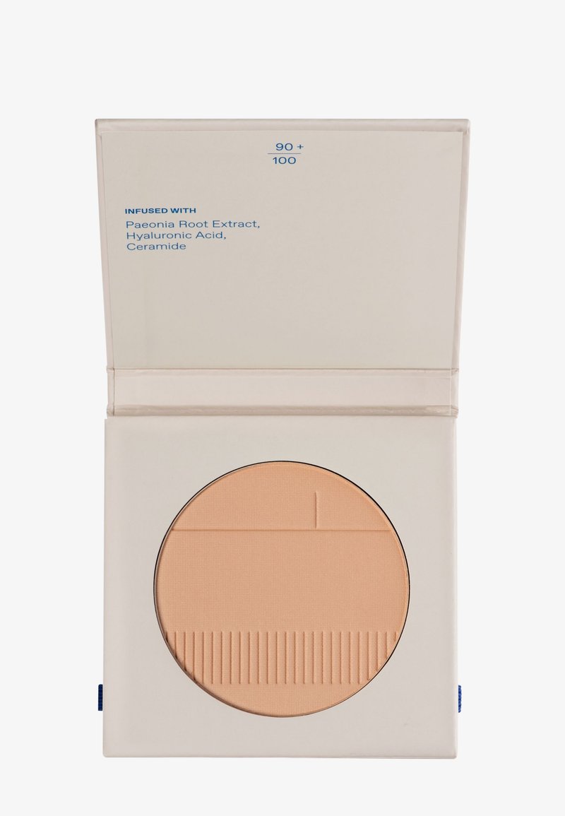 Poudre compacte dans un étui carré. Couleur beige clair avec une surface texturée. Étiquette à l'intérieur indiquant les ingrédients en texte bleu.
