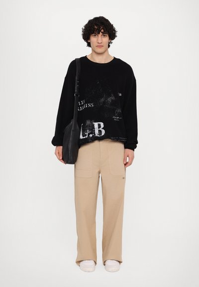 Sweatshirt noir surdimensionné avec imprimés graphiques blancs, associé à un pantalon cargo beige doté de poches, et des baskets blanches. Sac à épaule noir inclus.