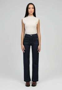 Femme se tenant face à l'avant, portant un haut sans manches blanc, un jean taille haute sombre avec des poches avant et des bottes à chevilles marron, sur un fond gris.