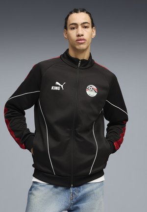 Młody mężczyzna z zaplecionymi włosami, ubrany w czarną kurtkę na zamek z czerwonymi akcentami i sportowymi logo, stojący z rękami w kieszeniach na szarym tle.