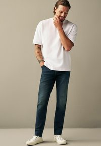 Tricou alb cu mânecă scurtă, croială lejeră, asortat cu blugi decolorati, bleumarin. Adidași albi completează ținuta, stând în fața unui fundal neutru.