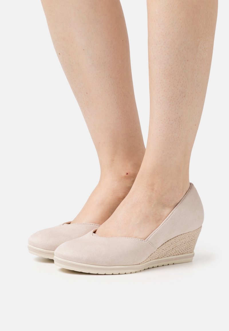 Pieds portant des chaussures compensées beige clair avec des côtés texturés et des bouts arrondis, présentés sur un fond blanc uni.