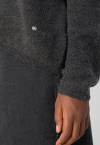 Manchette d'un pull gris foncé en tissu pelucheux, retroussée au poignet avec une petite étiquette métallique MOS MOSHI, associée à un pantalon en jean gris foncé et une main visible.