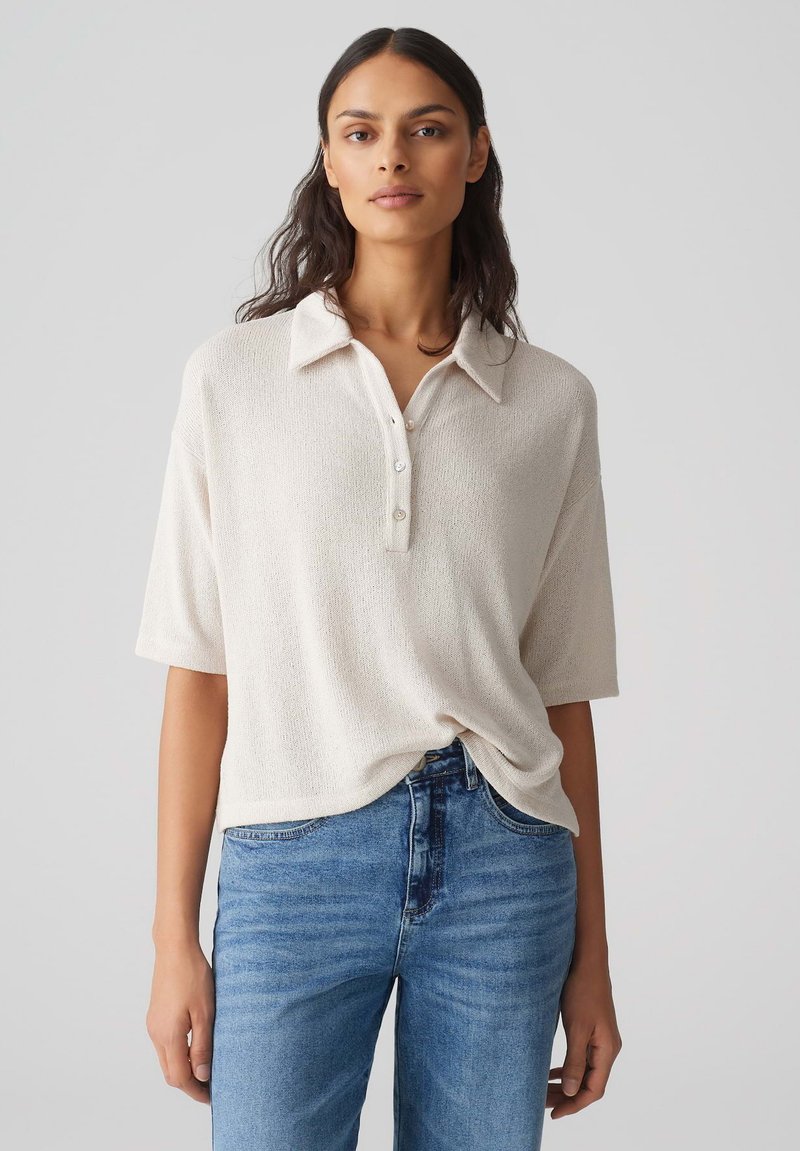 Helle beige Polohemd aus strukturiertem Stoff, mit kurzen Ärmeln und Knopfleiste. Kombiniert mit mitteltiefblauen, hochgeschnittenen Jeans.