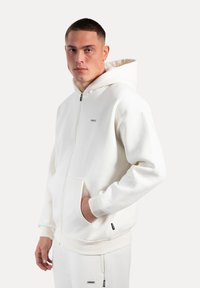 Hvid zip-hoodie lavet af blødt stof med en stor hætte, frontlommer, ribbede manchetter og en lille logo-detail på brystet.