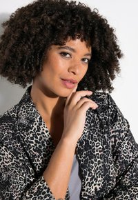 Blazer con estampado de leopardo en una combinación de colores negro y beige, con solapas muestradas y un corte relajado. La modelo posa con la mano cerca del mentón.