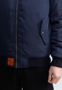 Marineblauwe bomberjack met een ritssluiting aan de voorkant, geribde manchetten, zijzak met drukknop en een oranje merklabel op de onderste zoom.