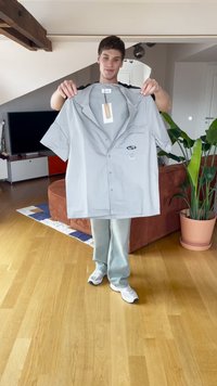 Joven en interiores sosteniendo una camisa de botones de manga corta de color gris claro con un bolsillo en el pecho y etiqueta visible.