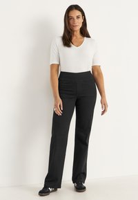 Nainen seisoo valkoista taustaa vasten, päällään lyhythihainen valkoinen toppi, korkeavyötäröiset mustat housut ja mustat lenkkarit, joissa on valkoiset raidat.