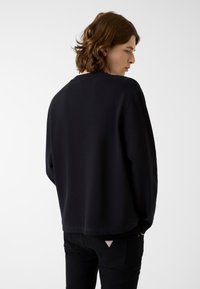 Czarna bluza z prążkowaną fakturą i okrągłym dekoltem, noszona z czarnymi spodniami, które mają małą czerwoną metkę z logo na tylnej kieszeni.