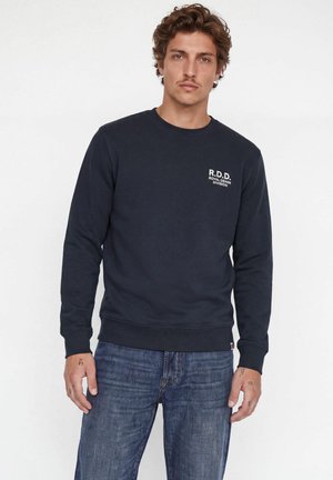 Mann mit hellbraunen lockigen Haaren, der einen schwarzen Pullover mit dem Text "R.D.D Royal Denim Division" und blaue Jeans trägt und vor einem weißen Hintergrund steht.