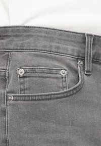 Κοντινό πλάνο τσέπης γκρι τζιν με μεταλλικά καρφιά και θηλιά για ζώνη, σε συνδυασμό με λευκό ριμπ ύφασμα τοπ.