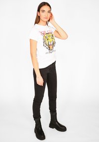 Ung kvinde står op, iført en t-shirt med et hvidt tiger-graphic, sorte stramme jeans og sorte klodsede støvler, med en hånd der rører ved hendes hår.