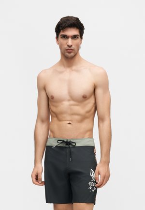 Νεαρός άνδρας χωρίς πουκάμισο με κοντά σκούρα μαλλιά, που φοράει μαύρο και γκρι μαγιό με λευκό γραφικό σχέδιο στο αριστερό πόδι.