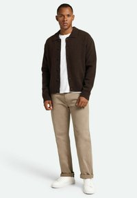 Cárdigan marrón de punto con cuello, con cremallera frontal, combinado con una camiseta blanca y pantalones beige, accessorios con zapatillas blancas.