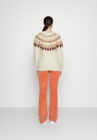 Pull crème en tricot avec un motif géométrique marron et beige sur le plastron, associé à un pantalon évasé en velours côtelé orange, vu de dos.