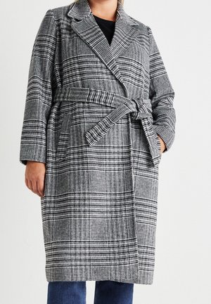 Femme portant un manteau long à carreaux noir et blanc ceinturé, avec de larges revers, sur un jean bleu et un haut noir.