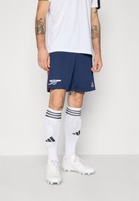 Marineblauwe voetbalkoeken met een wit logo en rode accenten, gecombineerd met knie-hoge witte sokken met zwarte strepen en patroondetails, en witte voetbalschoenen.