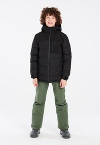 Doudoune noire avec capuche, fermeture éclair et détails de poches avant, associée à un pantalon de neige vert et des bottes en caoutchouc noires.