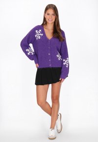 Cardigan lila din lână, cu decolteu în V, nasturi pe față și modele de fulgi de zăpadă albi pe mâneci. Asortat cu o fustă neagră plisată.