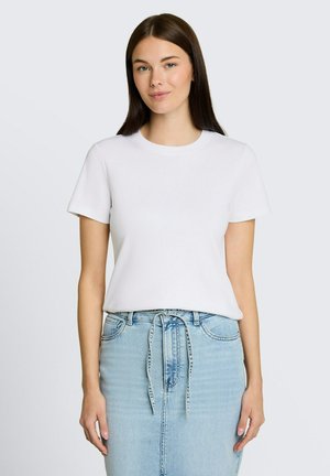 Kobieta z długimi brązowymi włosami, ubrana w gładki biały t-shirt z krótkim rękawem i jasnoniebieską spódnicę dżinsową z przewiązanym paskiem, stojąca na jednolitym tle.