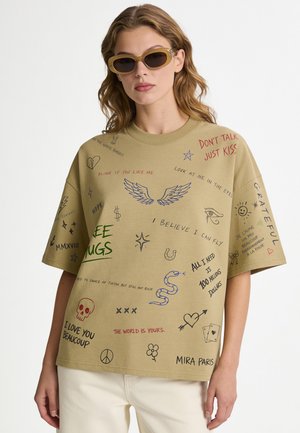 Vrouw draagt een oversized beige T-shirt met meerkleurige handgeschreven woorden, symbolen en tekeningen, gecombineerd met beige broek en zonnebril.
