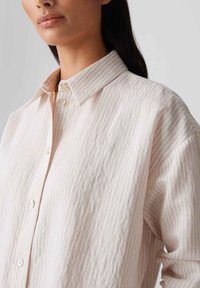 Lichtroze gestreepte knopshirt van zacht fabric. Beschikt over een klassieke kraag en een voorpaneel met knopen. Subtiele textuur en een ontspannen pasvorm.