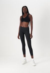 Black sports bra with a logo band and matching high-waisted leggings. Smooth, stretchy fabric. Model wears white sneakers.